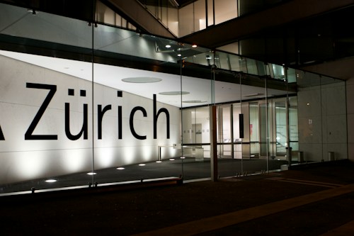 A modern building facade featuring large glass panels and a brightly illuminated interior. The word 'Z&uuml;rich' is displayed prominently across the glass. The interior appears minimalist and well-lit, with visible structural elements and reflections on the glass surface.
