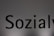 The image features a close-up of the word 'Sozialy' printed in bold black letters on a plain, light gray background. The lighting creates a subtle gradient effect on the background.