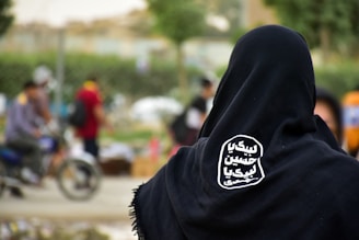A person wearing a black garment stands with their back to the camera. On the back of the garment, there's white Arabic script in a stylized oval shape. In the background, people are moving around, with a blurred focus, including someone on a motorbike. The setting appears to be an outdoor area, possibly a market or street scene.