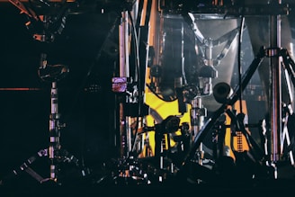 An intricate arrangement of metallic drum hardware. Chrome drum stands and hoops are illuminated by dim, moody lighting with a contrasting yellowish glow in the background.
