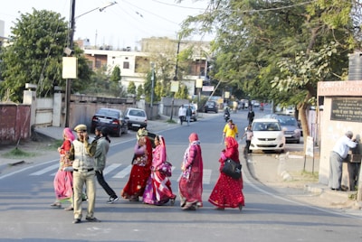 A group of people in traditional clothing are crossing a street in an urban setting. One person is wearing a uniform, possibly of a security or police officer. Vehicles are parked along the roadside, and trees and buildings are visible in the background.