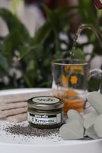 A jar labeled in Cyrillic script indicating Matcha cream honey is placed on a surface surrounded by black chia seeds. A glass mug decorated with yellow designs contains a light brown liquid, being filled from a spout peeking from above. Behind the items, green foliage creates a natural backdrop with a well-lit setting that suggests freshness.