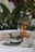 A jar labeled in Cyrillic script indicating Matcha cream honey is placed on a surface surrounded by black chia seeds. A glass mug decorated with yellow designs contains a light brown liquid, being filled from a spout peeking from above. Behind the items, green foliage creates a natural backdrop with a well-lit setting that suggests freshness.