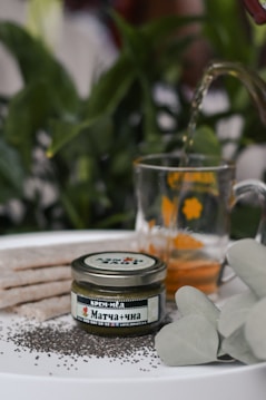 A jar labeled in Cyrillic script indicating Matcha cream honey is placed on a surface surrounded by black chia seeds. A glass mug decorated with yellow designs contains a light brown liquid, being filled from a spout peeking from above. Behind the items, green foliage creates a natural backdrop with a well-lit setting that suggests freshness.