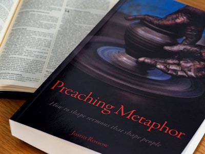 An open book with text is positioned next to another book titled 'Preaching Metaphor: How to shape sermons that shape people' by Justin Rossow. The cover features an image of hands shaping clay on a pottery wheel.