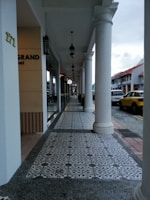 An outdoor walkway of a commercial area featuring decorative tiled flooring and white columns supporting an overhang. Hanging lamps align the path, leading the eye towards the distance. The side of a building displays partial signage, and a yellow vehicle is parked along the adjacent street.