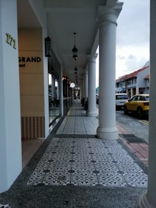 An outdoor walkway of a commercial area featuring decorative tiled flooring and white columns supporting an overhang. Hanging lamps align the path, leading the eye towards the distance. The side of a building displays partial signage, and a yellow vehicle is parked along the adjacent street.
