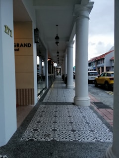 An outdoor walkway of a commercial area featuring decorative tiled flooring and white columns supporting an overhang. Hanging lamps align the path, leading the eye towards the distance. The side of a building displays partial signage, and a yellow vehicle is parked along the adjacent street.