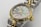 A luxury wristwatch featuring a mother-of-pearl dial adorned with diamond hour markers and gold accents. The watch has a two-tone design with a polished metal bracelet and a prominent gold bezel. The brand name and additional text are engraved on the dial.