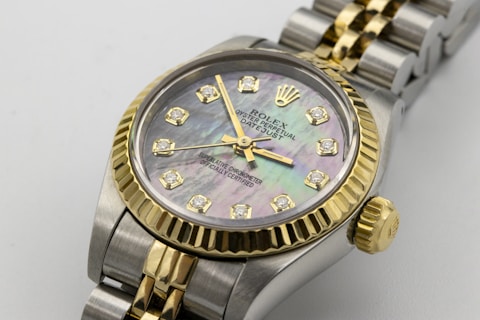 A luxury wristwatch featuring a mother-of-pearl dial adorned with diamond hour markers and gold accents. The watch has a two-tone design with a polished metal bracelet and a prominent gold bezel. The brand name and additional text are engraved on the dial.