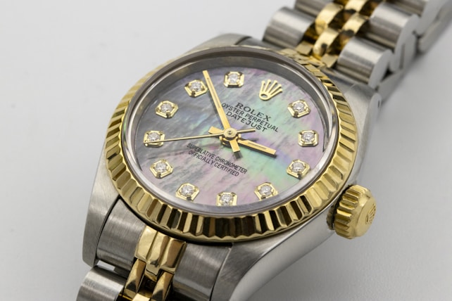 A luxury wristwatch featuring a mother-of-pearl dial adorned with diamond hour markers and gold accents. The watch has a two-tone design with a polished metal bracelet and a prominent gold bezel. The brand name and additional text are engraved on the dial.