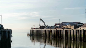 A serene waterfront scene with a dock stretching into calm waters. The sky is clear with a few clouds. There is a structure with a sloped roof labeled 'RESTAURANT' and a construction crane is visible. Piles of materials and equipment are on the dock, which is reflected in the water.