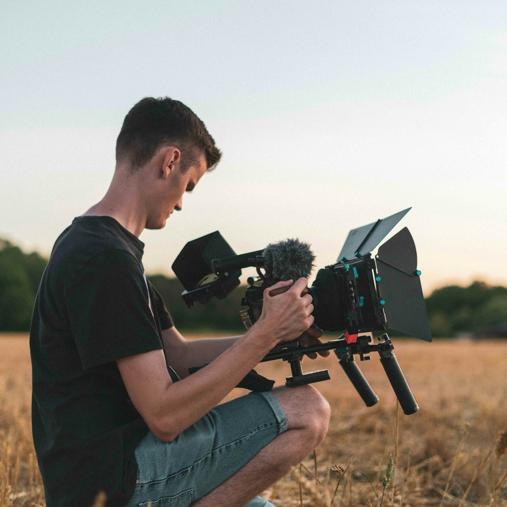 man in black t-shirt and blue denim shorts sitting on brown grass field holding black