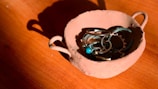 A rustic, handcrafted clay bowl containing a collection of silver jewelry, including rings and bracelets with turquoise stones, placed on a wooden surface. The lighting casts sharp shadows and highlights the texture of the clay.