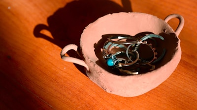 A rustic, handcrafted clay bowl containing a collection of silver jewelry, including rings and bracelets with turquoise stones, placed on a wooden surface. The lighting casts sharp shadows and highlights the texture of the clay.