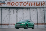 A bright green car is parked in front of a large, industrial-style building with a textured surface. Above the car is a large sign with Cyrillic text in red and white. The car's sleek design contrasts with the stark, geometric patterns of the building behind it.