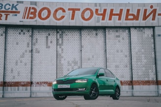 A bright green car is parked in front of a large, industrial-style building with a textured surface. Above the car is a large sign with Cyrillic text in red and white. The car's sleek design contrasts with the stark, geometric patterns of the building behind it.