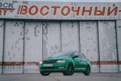 A bright green car is parked in front of a large, industrial-style building with a textured surface. Above the car is a large sign with Cyrillic text in red and white. The car's sleek design contrasts with the stark, geometric patterns of the building behind it.
