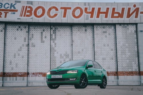 A bright green car is parked in front of a large, industrial-style building with a textured surface. Above the car is a large sign with Cyrillic text in red and white. The car's sleek design contrasts with the stark, geometric patterns of the building behind it.