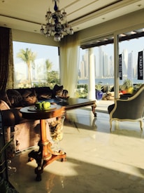 A luxurious living room with a classic wooden table set with green apples, ornate furniture including a velvet sofa and elegant chair. Large glass windows provide a view of a city skyline, palm trees, and a body of water, bathed in warm sunlight. A decorative chandelier hangs from the ceiling.
