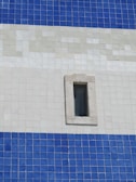 An exterior wall composed of square tiles in shades of blue and white, featuring a small, rectangular window with a stone frame. The tile pattern is organized in horizontal stripes with distinct color transitions.