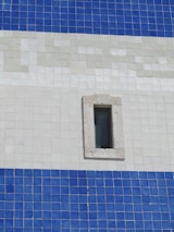 An exterior wall composed of square tiles in shades of blue and white, featuring a small, rectangular window with a stone frame. The tile pattern is organized in horizontal stripes with distinct color transitions.