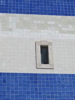 An exterior wall composed of square tiles in shades of blue and white, featuring a small, rectangular window with a stone frame. The tile pattern is organized in horizontal stripes with distinct color transitions.