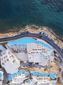 An aerial view of a coastal resort with a series of interconnected white buildings and blue swimming pools. The buildings are arranged in a complex layout, overlooking a road that runs parallel to the shoreline. The sea is visible, with its deep blue and green hues contrasting with the white architecture and the lighter blue of the pools.