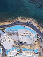An aerial view of a coastal resort with a series of interconnected white buildings and blue swimming pools. The buildings are arranged in a complex layout, overlooking a road that runs parallel to the shoreline. The sea is visible, with its deep blue and green hues contrasting with the white architecture and the lighter blue of the pools.