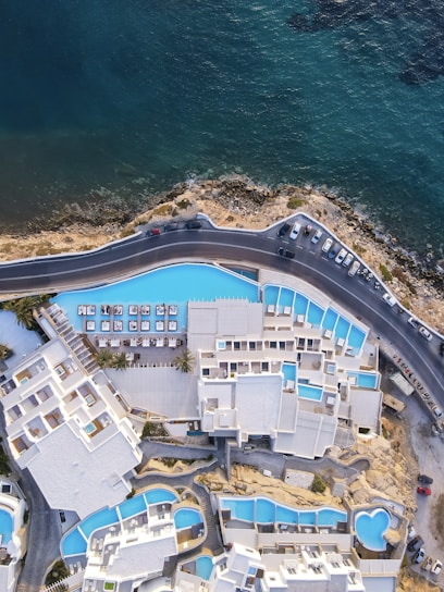 An aerial view of a coastal resort with a series of interconnected white buildings and blue swimming pools. The buildings are arranged in a complex layout, overlooking a road that runs parallel to the shoreline. The sea is visible, with its deep blue and green hues contrasting with the white architecture and the lighter blue of the pools.