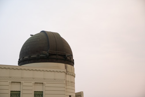 An observatory with a large, dark-colored dome sits atop a classical-style building. The building features intricate detailing and multiple rectangular windows. The sky appears overcast.