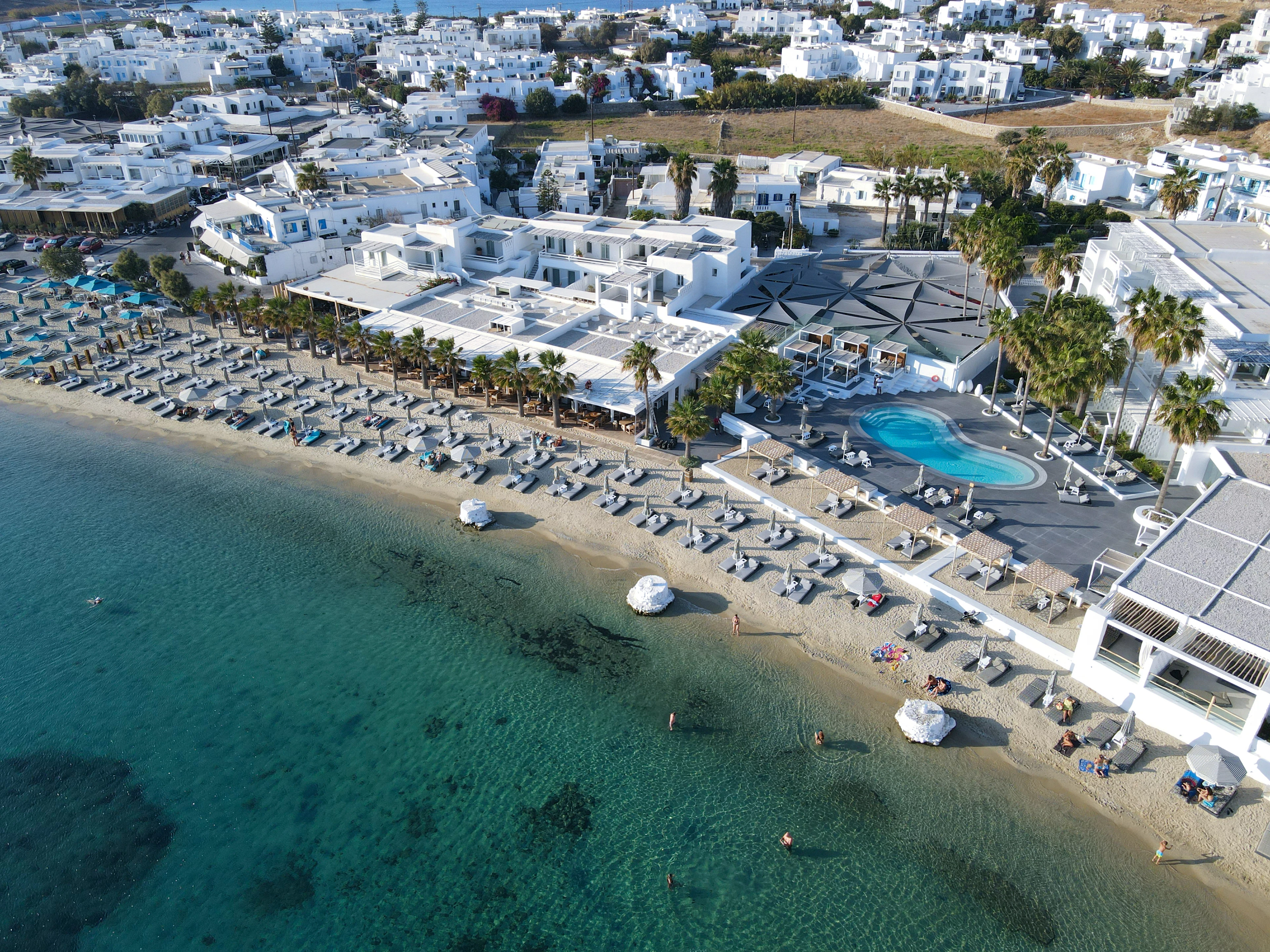 An aerial view of a Mykonos beach at golden hour: long ranks of straw parasols and loungers along the pale sand, whitewashed buildings and palm trees behind, and clear turquoise shallows in the foreground.