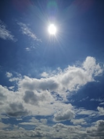 The image depicts a bright sun shining in a clear blue sky, with its rays extending outward. Fluffy white clouds are scattered across the sky, adding texture and depth to the scene.