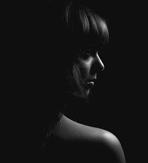 A black and white portrait featuring a person in profile view with soft lighting emphasizing facial features and shoulder against a dark background.