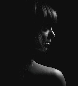 A black and white portrait featuring a person in profile view with soft lighting emphasizing facial features and shoulder against a dark background.