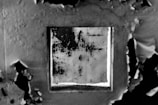A close-up view of a deteriorating wall with peeling paint surrounding a square patch of darker, textured surface. The contrasting textures and tones create a striking visual effect with remnants of the original paint hugging the edges of the square.