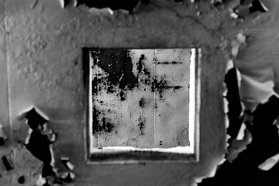 A close-up view of a deteriorating wall with peeling paint surrounding a square patch of darker, textured surface. The contrasting textures and tones create a striking visual effect with remnants of the original paint hugging the edges of the square.