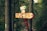 A vibrant path marker in a dense forest. The signpost features several directions, with a collage of colorful stickers and icons on orange and red backgrounds. Surrounding the sign are tall, thick trees with lush green foliage and a narrow trail winding through the forest. The area appears serene and natural.