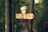 A vibrant path marker in a dense forest. The signpost features several directions, with a collage of colorful stickers and icons on orange and red backgrounds. Surrounding the sign are tall, thick trees with lush green foliage and a narrow trail winding through the forest. The area appears serene and natural.
