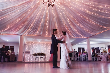 A bride and groom elegantly dancing under a beautifully draped ceiling adorned with warm, twinkling lights. Guests are seated at tables decorated with floral arrangements in the background, creating a romantic and festive atmosphere.