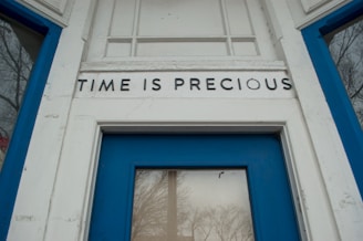 A close-up view of a doorway with a blue frame and the words 'TIME IS PRECIOUS' displayed above the door. The door window reflects bare tree branches.