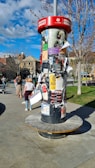 A cylindrical outdoor bulletin board is covered with various posters and flyers. It is set in a public area with a few people walking nearby. Trees with sparse branches and a building with arched windows are visible in the background. The sky is partly cloudy, allowing sunlight to cast shadows.