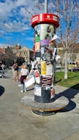 A cylindrical outdoor bulletin board is covered with various posters and flyers. It is set in a public area with a few people walking nearby. Trees with sparse branches and a building with arched windows are visible in the background. The sky is partly cloudy, allowing sunlight to cast shadows.