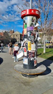 A cylindrical outdoor bulletin board is covered with various posters and flyers. It is set in a public area with a few people walking nearby. Trees with sparse branches and a building with arched windows are visible in the background. The sky is partly cloudy, allowing sunlight to cast shadows.