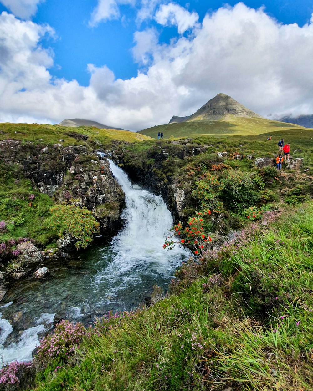 people standing on green grass field near waterfalls under blue and white cloudy sky during daytime