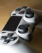 A close-up view of a video game controller featuring a sleek design with multiple buttons including action buttons with symbols, directional pad, and analog sticks. The controller is predominantly gray with black and colorful button details.