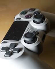 A close-up view of a video game controller featuring a sleek design with multiple buttons including action buttons with symbols, directional pad, and analog sticks. The controller is predominantly gray with black and colorful button details.