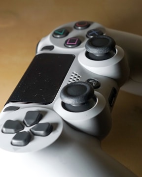 A close-up view of a video game controller featuring a sleek design with multiple buttons including action buttons with symbols, directional pad, and analog sticks. The controller is predominantly gray with black and colorful button details.