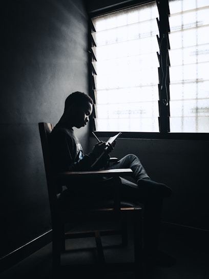 A person sitting on a chair near a window, reading or writing in a notebook. The scene is dimly lit, creating a silhouette effect against the bright window.