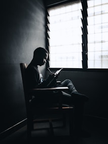 A person sitting on a chair near a window, reading or writing in a notebook. The scene is dimly lit, creating a silhouette effect against the bright window.